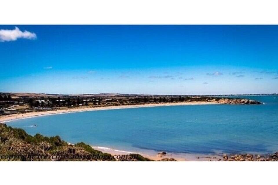 A coastal landscape with a clear blue sea, sandy beach, and a distant shoreline with buildings.
