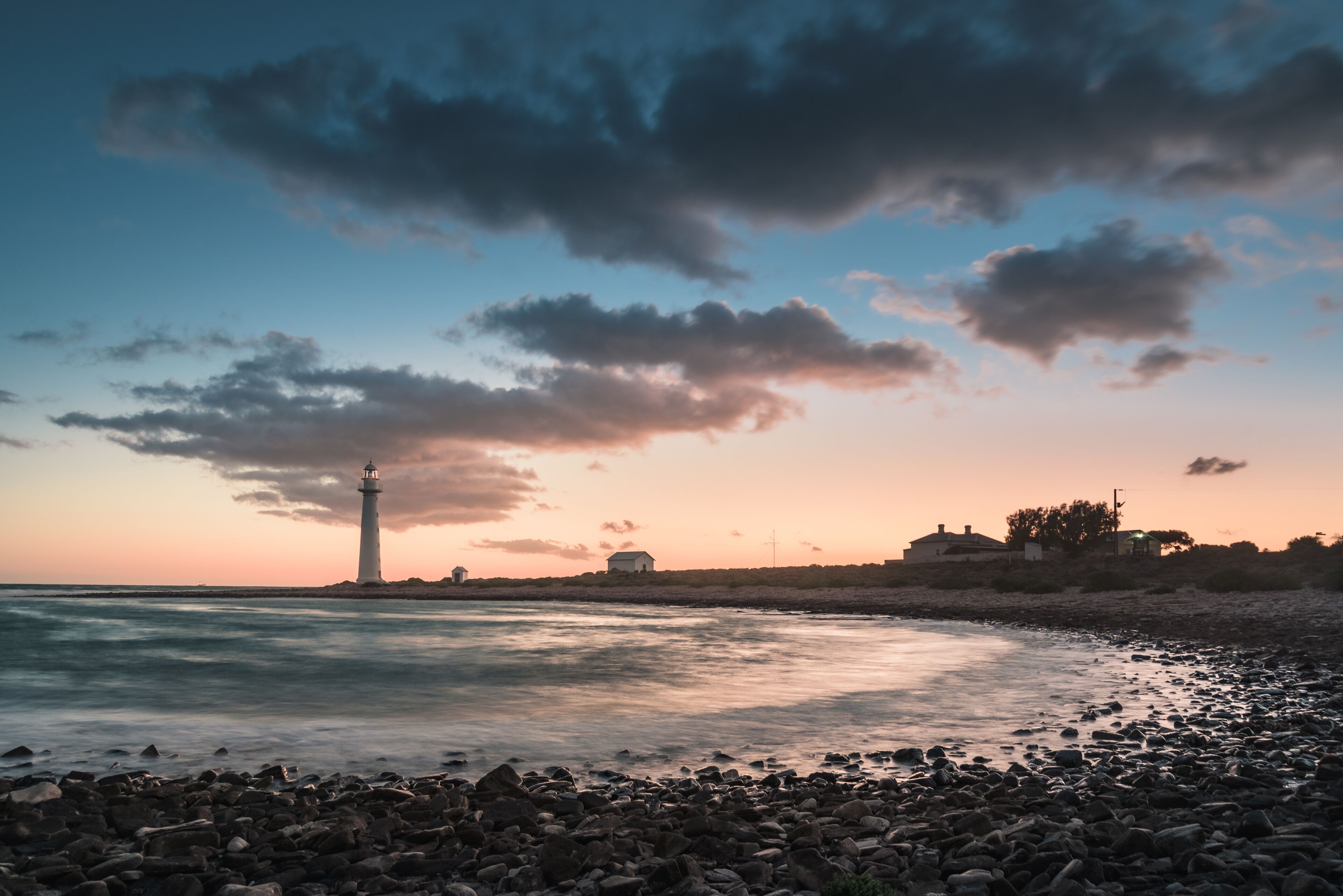 A rocky shoreline with a lighthouse, calm sea, and a sunset sky.