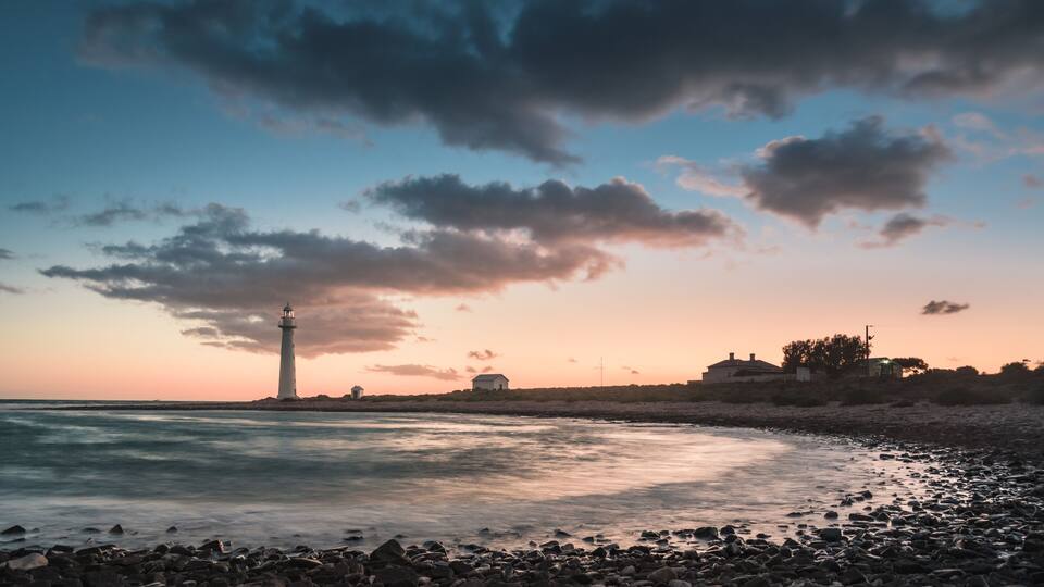 A rocky shoreline with a lighthouse, calm sea, and a sunset sky.