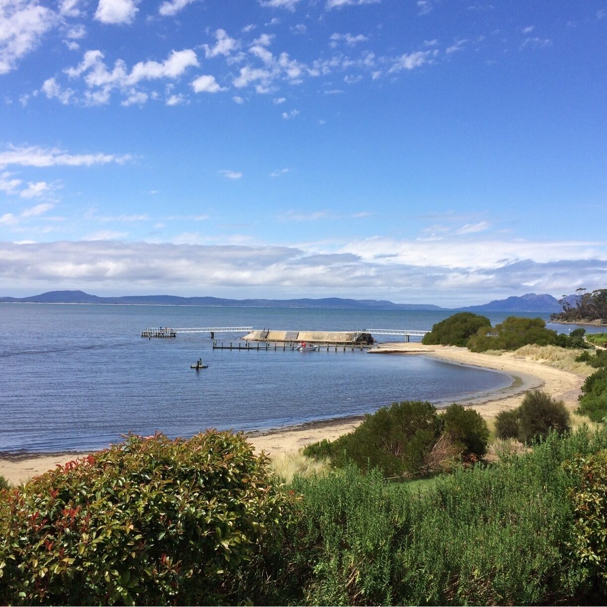 A coastal landscape with a sandy beach, a pier extending into the water, and distant mountains under a partly cloudy sky.