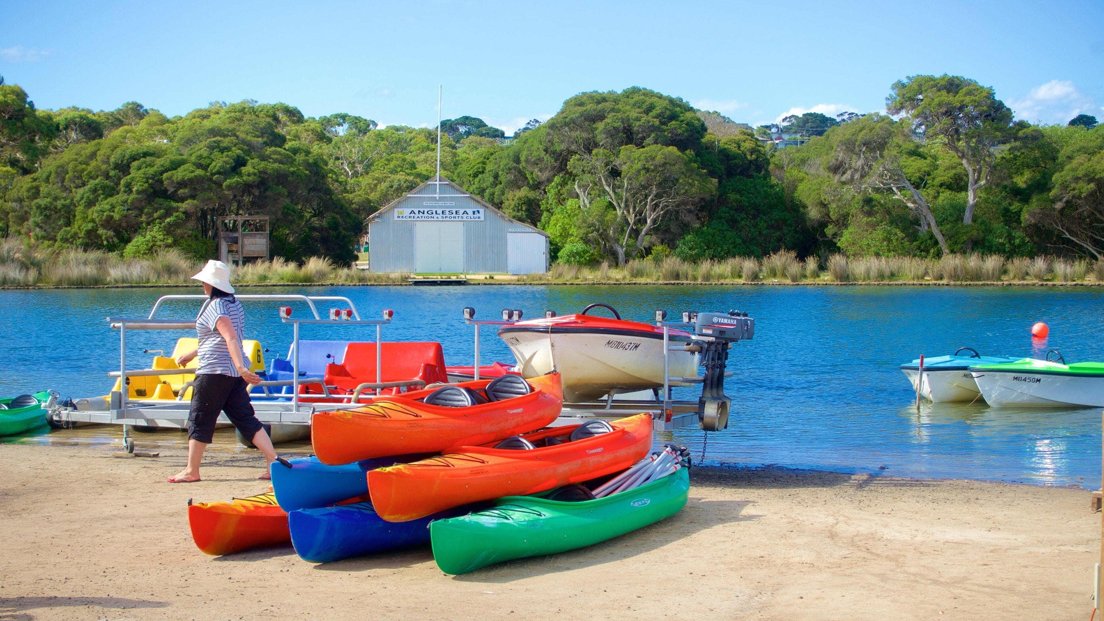 A sandy beach with several colorful kayaks and a motorboat docked near the water's edge. A person is walking away from the boats. In the background, there is a building with a sign that reads "ANGLESEA Recreation & Sports Club."
