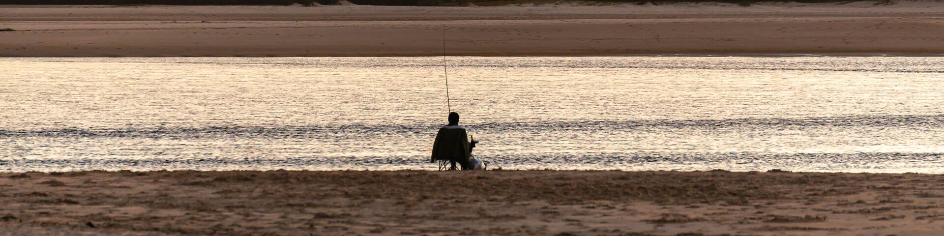 Rear View of Fisherman Sitting on the Beach at Sunset whit Mountain and Sea Landscape