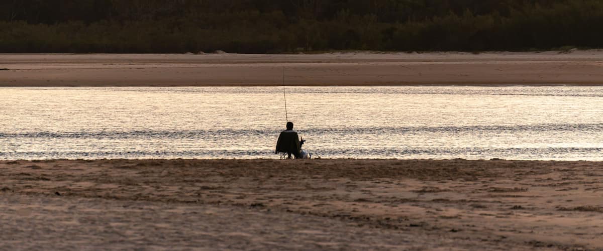 A person fishing by the water with a mountain in the background.