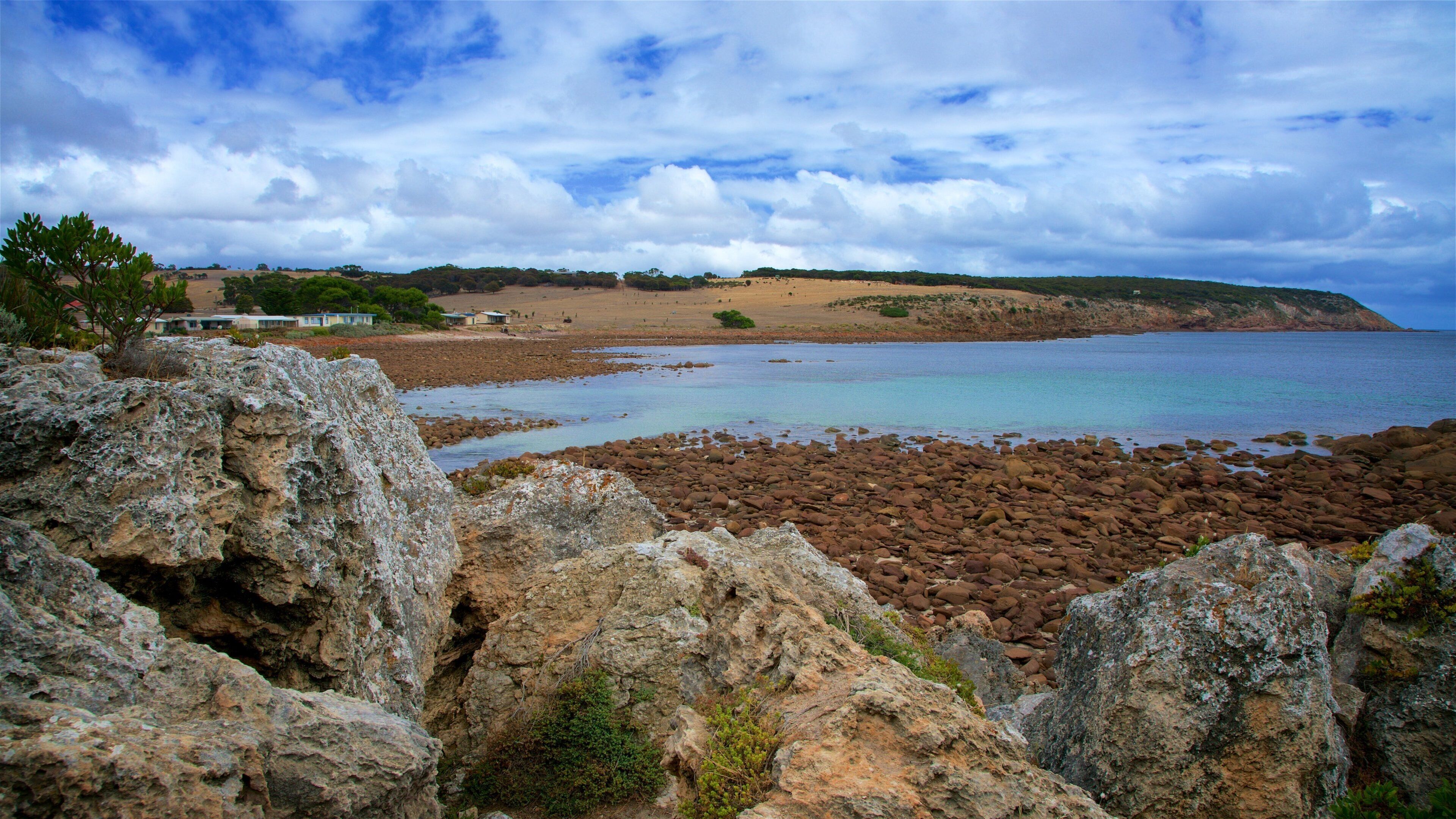 A rocky shoreline with a calm sea, a sandy beach, and a distant hill with sparse vegetation.