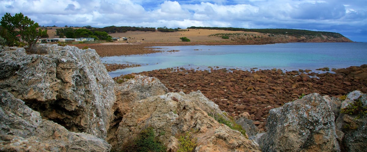 A rocky shoreline with a calm sea, a sandy beach, and a distant hill with sparse vegetation.
