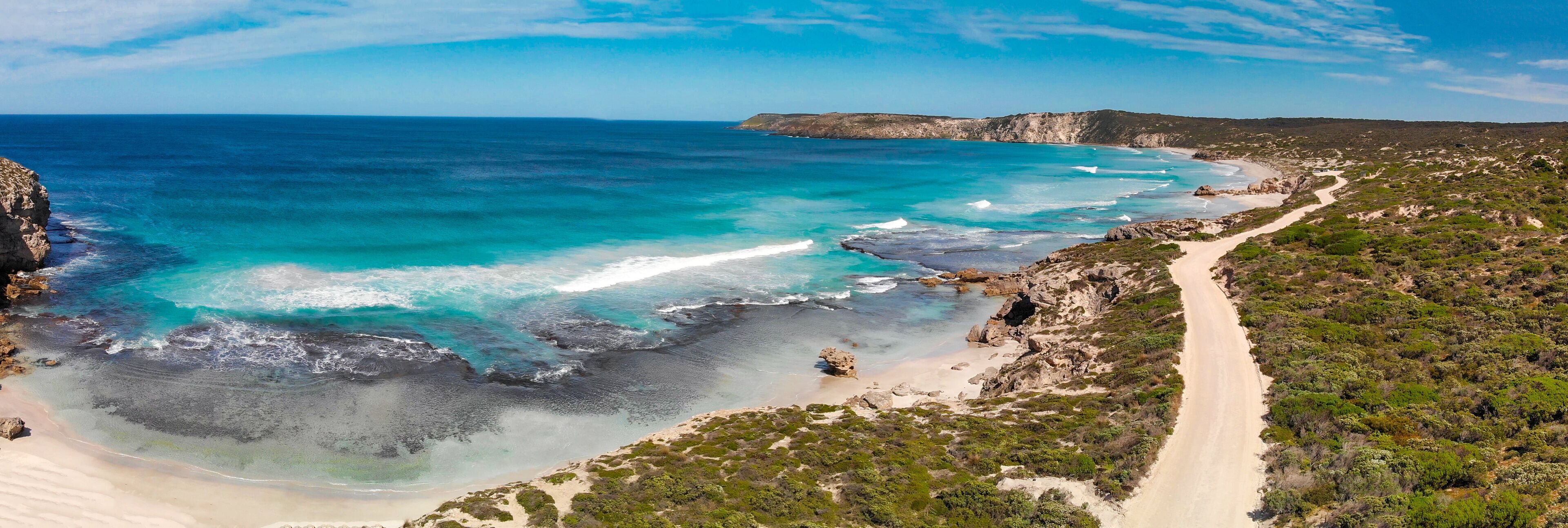 A coastal landscape with a sandy beach, clear turquoise waters, rocky cliffs, and a winding road.