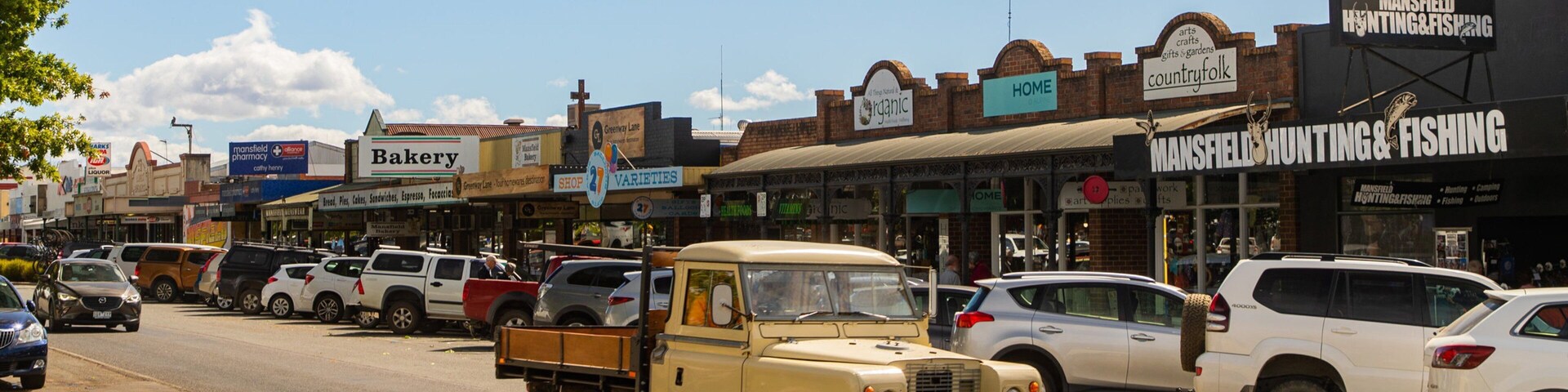 Una calle concurrida bordeada de diversos comercios, incluyendo una panadería, una tienda de artículos de caza y pesca, y una tienda de artesanía rural.