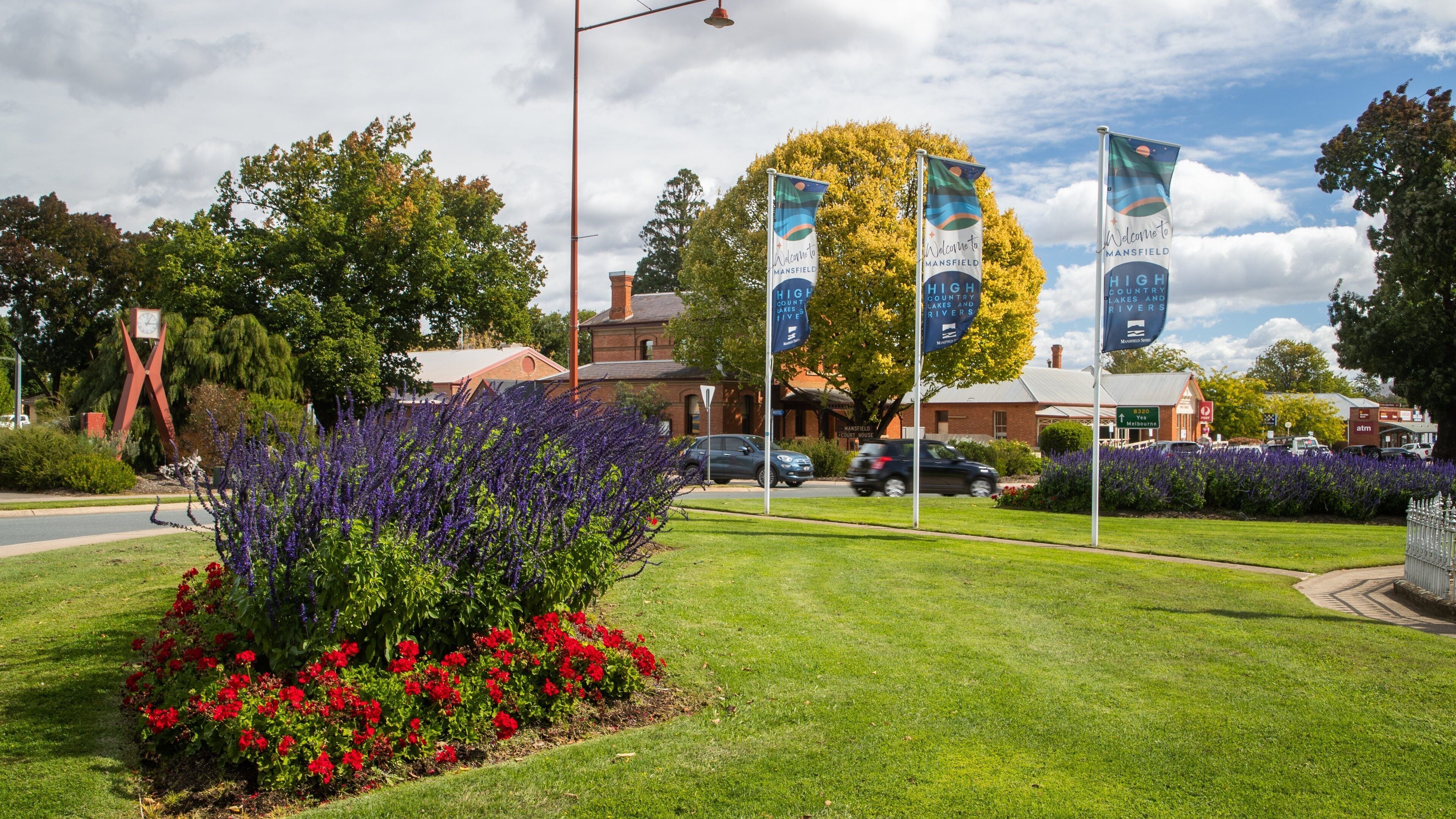 A well-maintained lawn with red flowers, a clock tower, and flags in a town square.