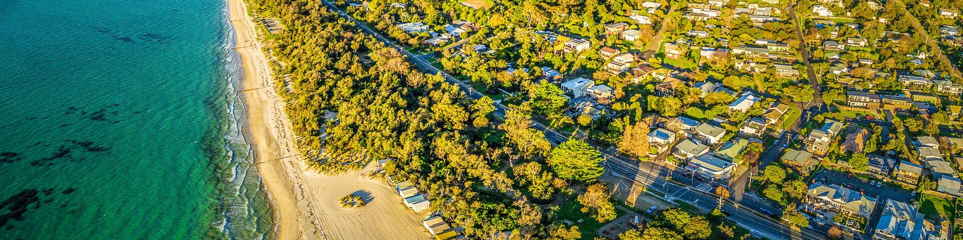 An aerial view of a coastal town with a beach, dense greenery, and a hilly backdrop.