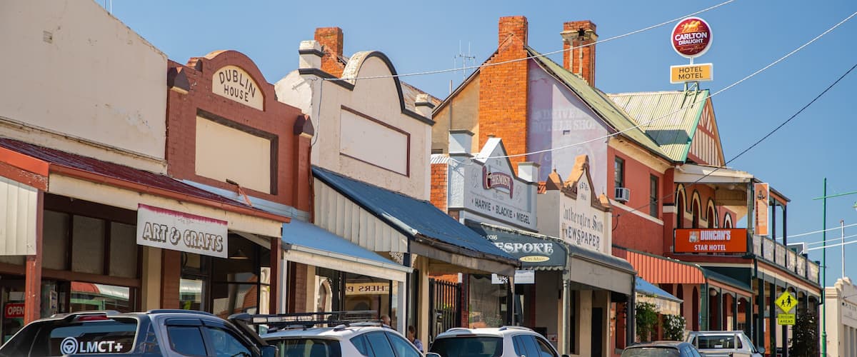 A street lined with shops including a hotel, a chemist, and a newspaper.