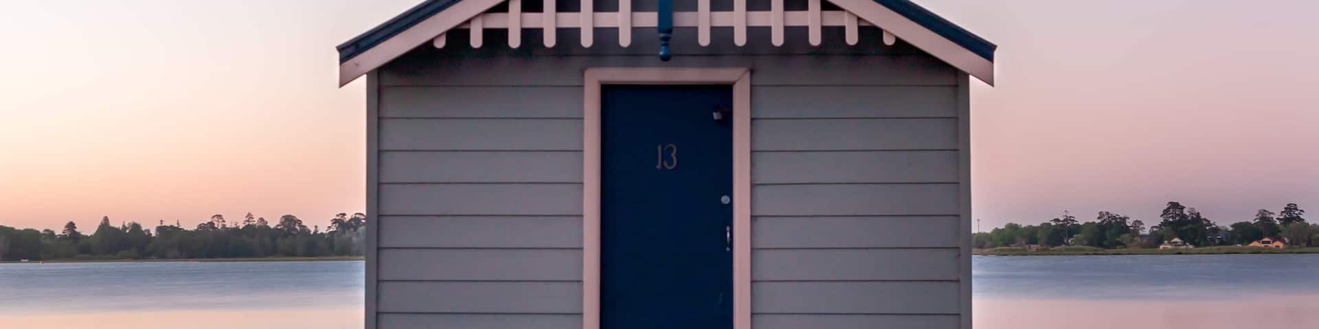 A small wooden boathouse with a blue door and a white trim, situated on a wooden dock extending into calm water.