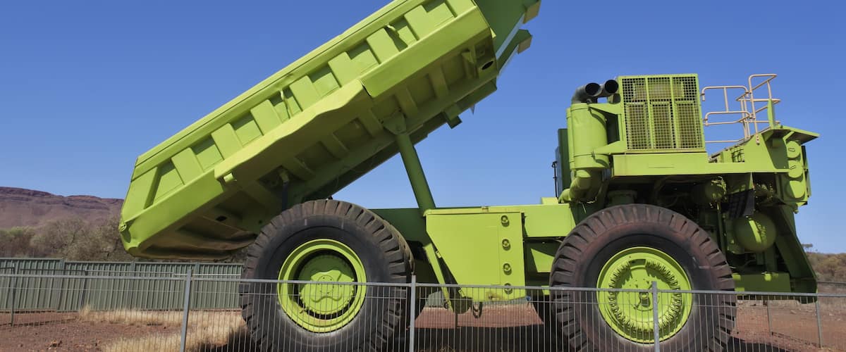 A large green agricultural vehicle with a raised bed, large tires, and a metal fence in the foreground.