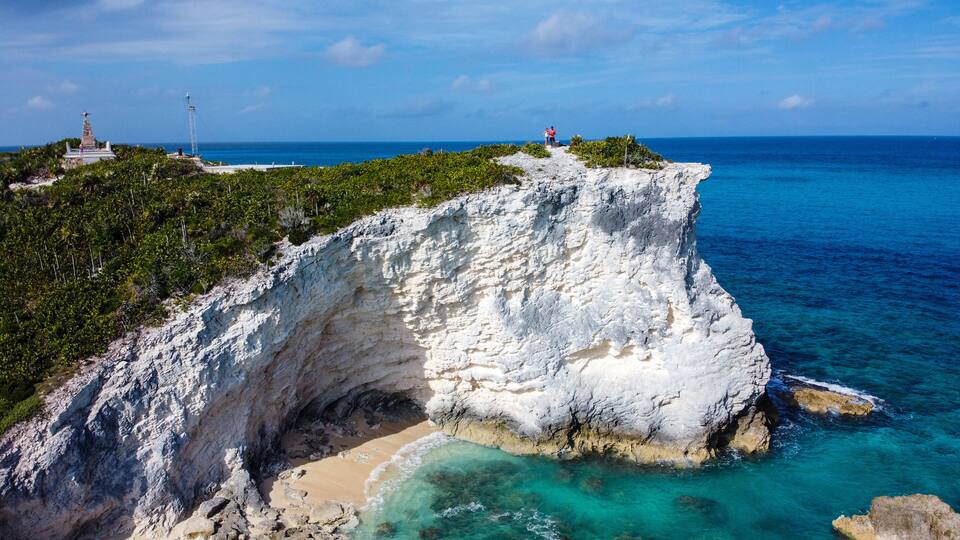 A white cliff with a beach at its base, a lighthouse, and a person standing on the cliff edge.