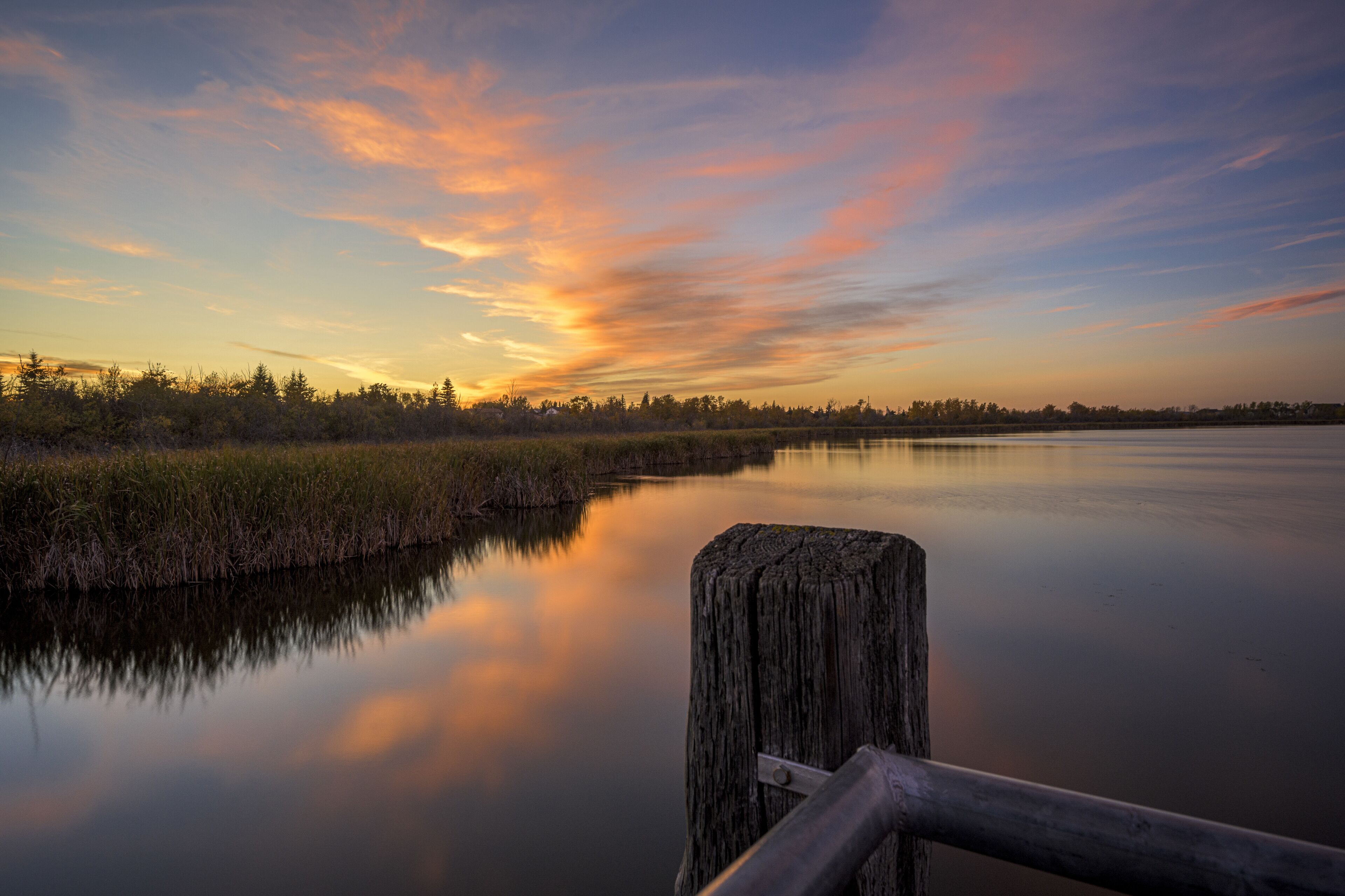 A calm river with a wooden post in the foreground, surrounded by reeds and trees under a sunset sky.
