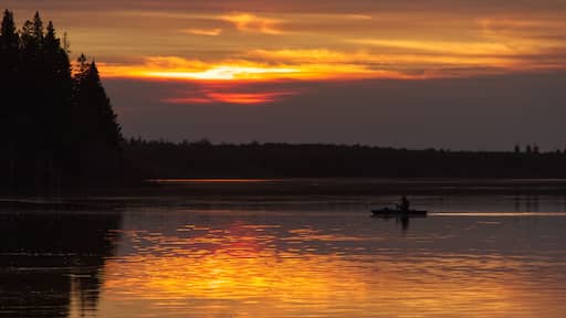 Un atardecer sobre un lago tranquilo, con la silueta de árboles a la izquierda y una persona en un bote a lo lejos.