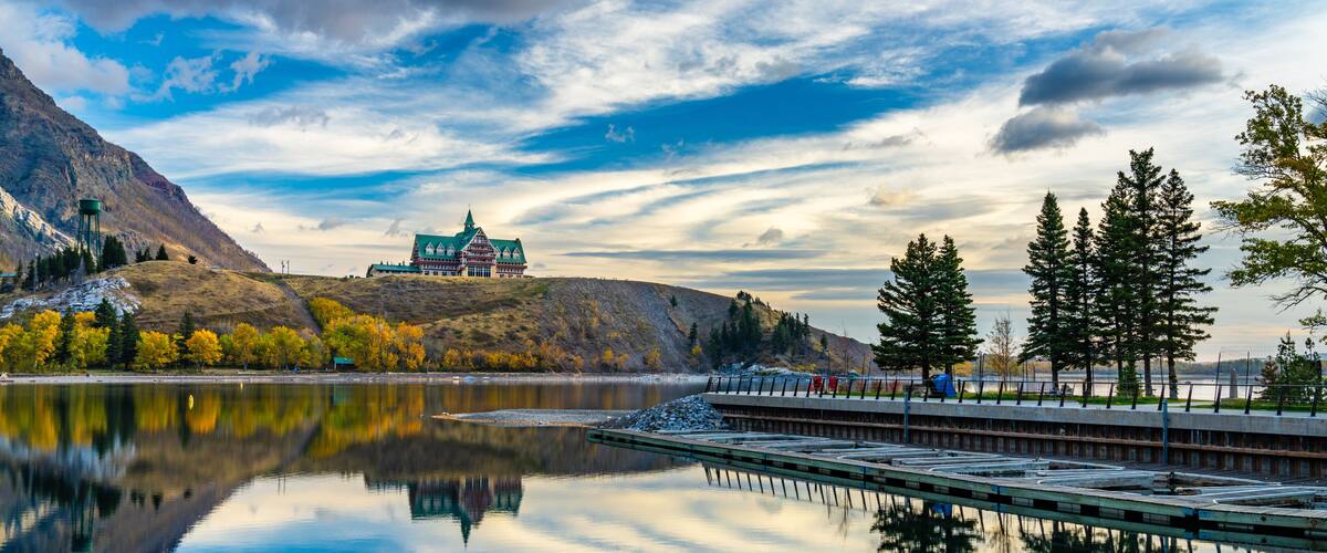 Waterton Lakes National Park lakeshore in autumn foliage season morning. Blue sky, colourful clouds reflect on lake surface like a mirror in sunrise. Fall color landscape. Landmarks in Alberta, Canada