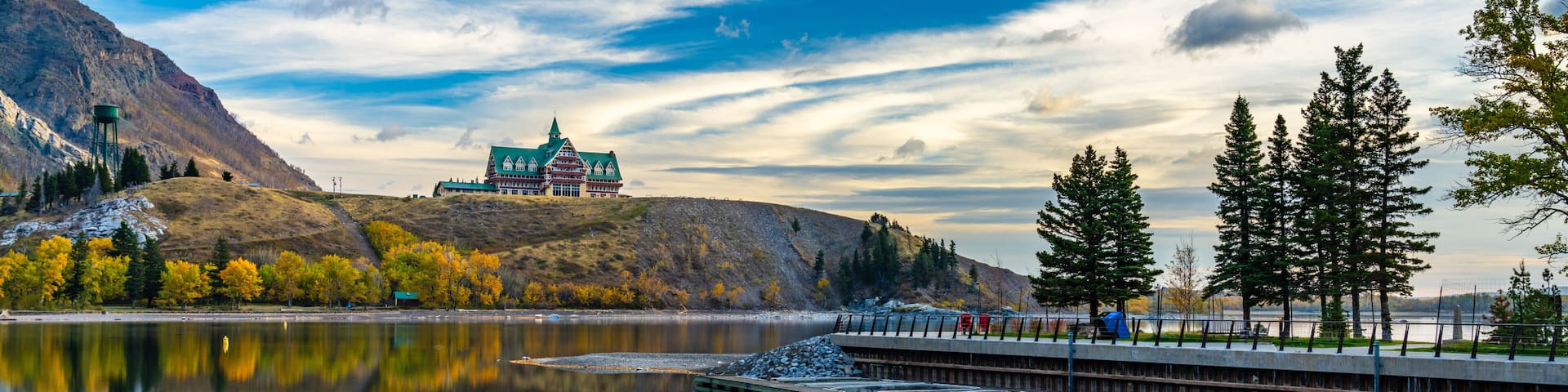 Waterton Lakes National Park lakeshore in autumn foliage season morning. Blue sky, colourful clouds reflect on lake surface like a mirror in sunrise. Fall color landscape. Landmarks in Alberta, Canada