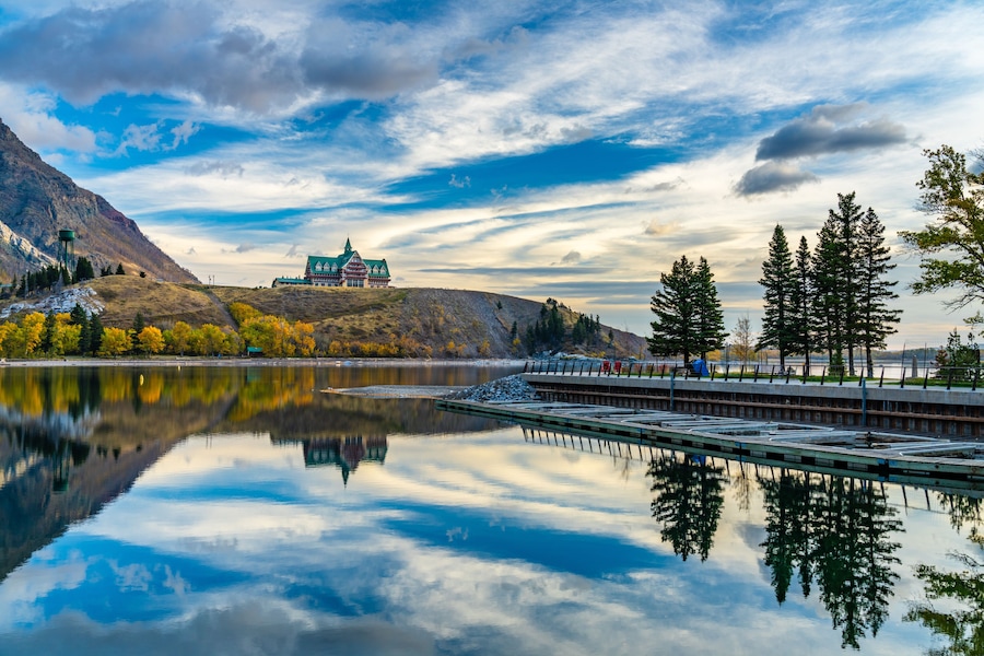 Waterton Lakes National Park lakeshore in autumn foliage season morning. Blue sky, colourful clouds reflect on lake surface like a mirror in sunrise. Fall color landscape. Landmarks in Alberta, Canada