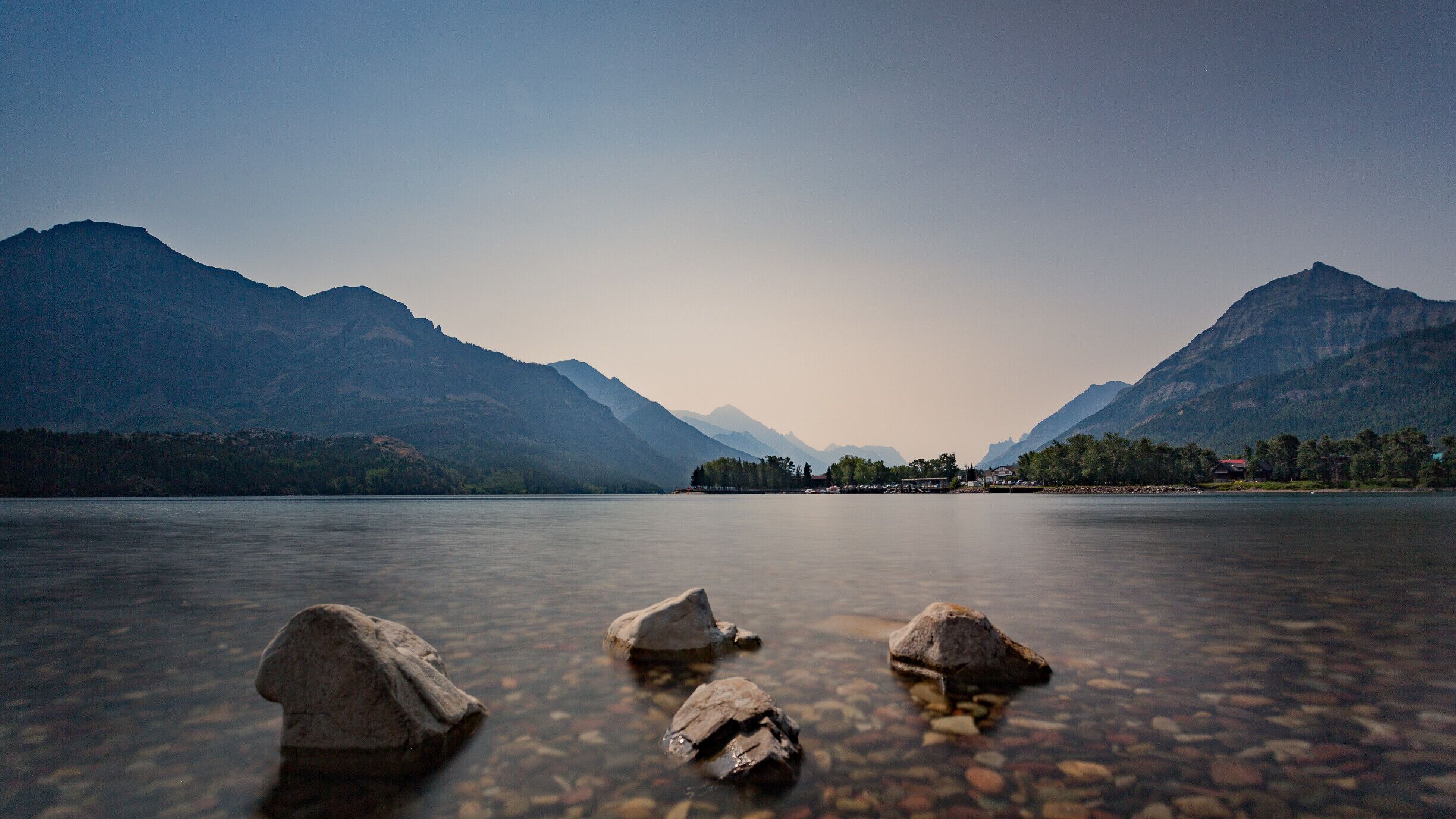 A calm lake with clear water, surrounded by mountains and a small settlement with houses.
