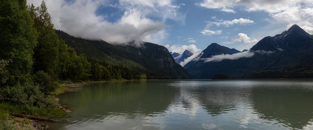 A calm lake surrounded by dense forest and mountains with snow-capped peaks.