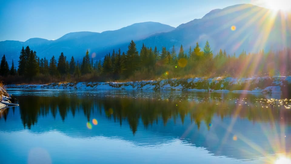A calm lake reflecting trees and mountains under a clear sky.