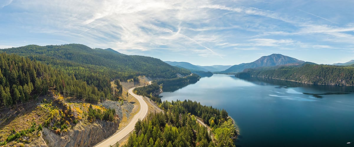A winding road through a forested valley leading to a calm lake surrounded by mountains.