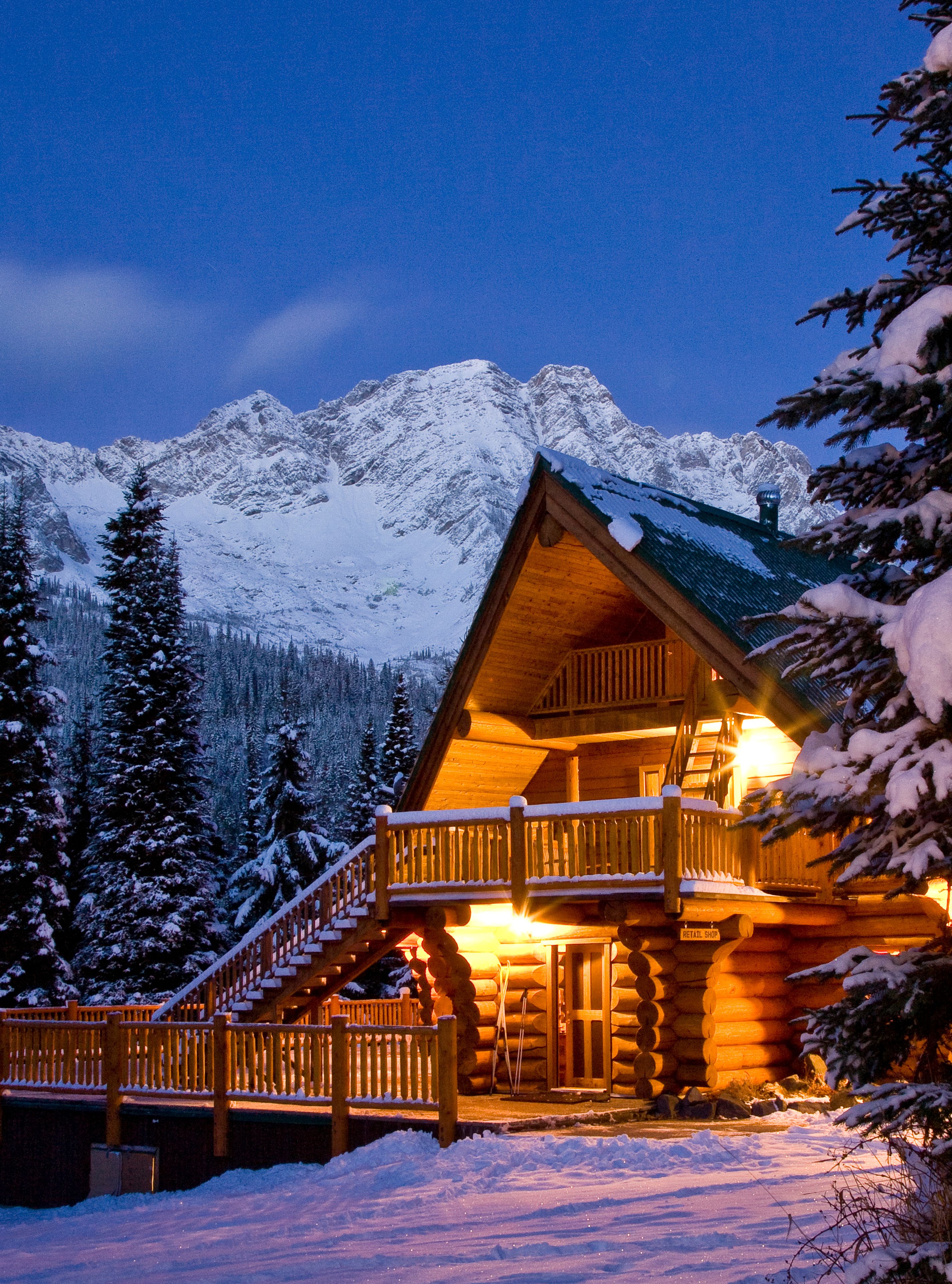 A wooden cabin with a balcony, surrounded by snow-covered trees and mountains in the background.