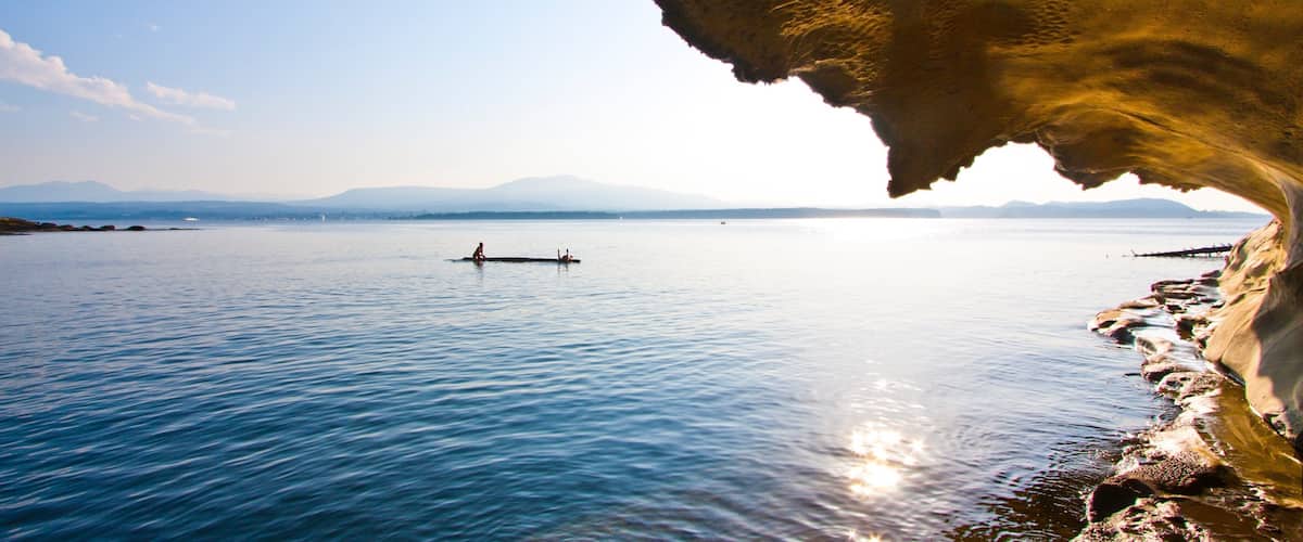 A calm lake with a person rowing a boat, surrounded by mountains and a rocky shoreline.