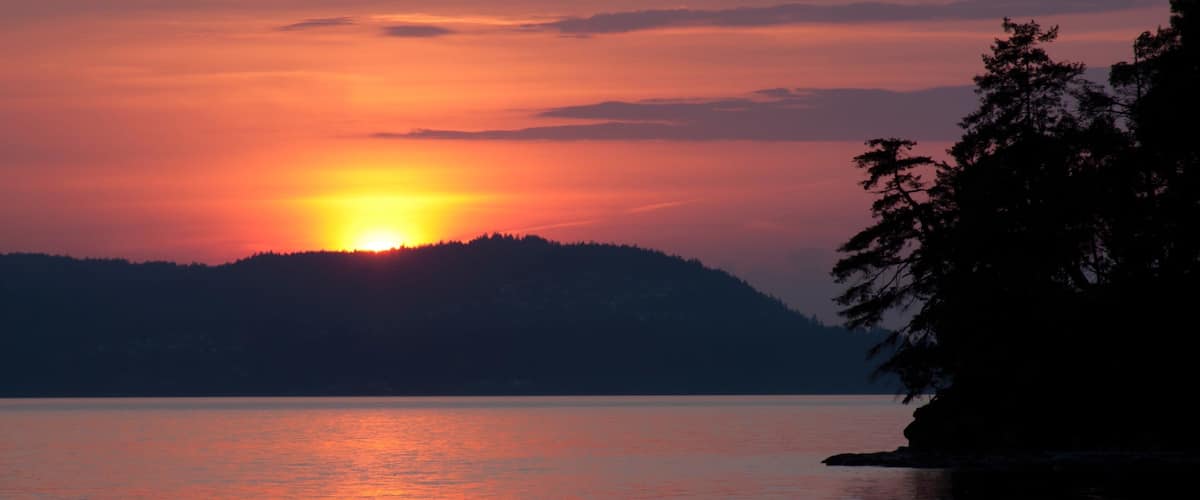 A sunset over a calm lake with a silhouette of a forested hill in the background.