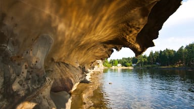 Formação rochosa natural com uma abertura semelhante a uma caverna sobre um lago calmo, rodeada por árvores e uma casa distante.