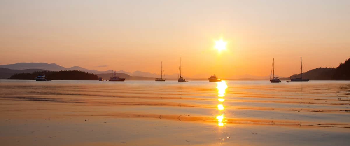 A sunset over a calm lake with several boats and a distant mountain range.