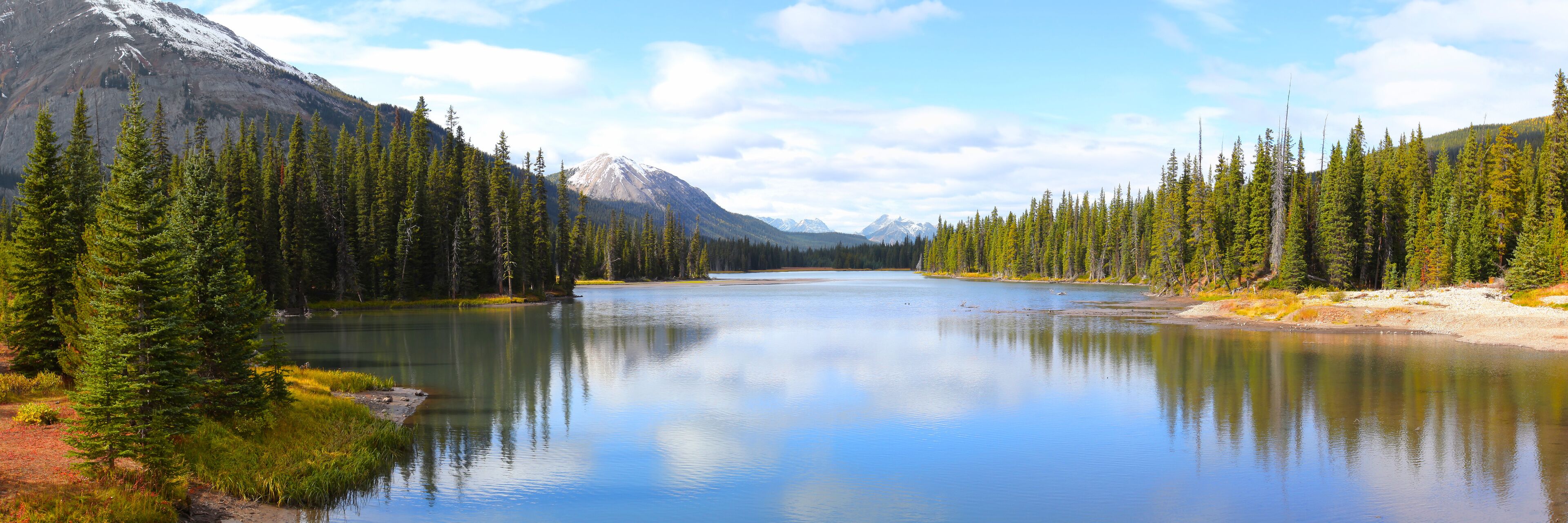 A calm lake surrounded by dense coniferous forest and snow-capped mountains.