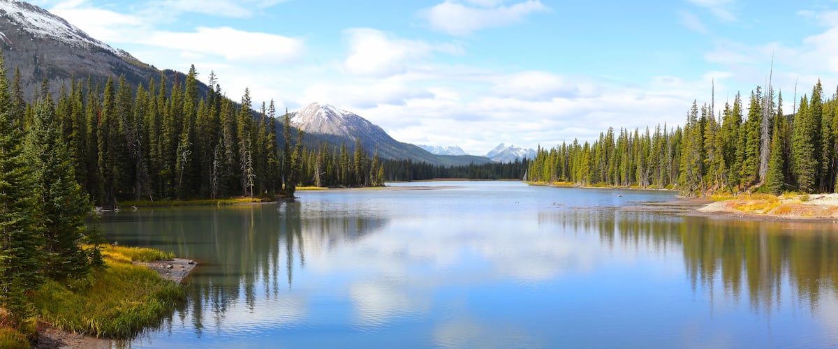 A calm lake surrounded by dense coniferous forest and snow-capped mountains.