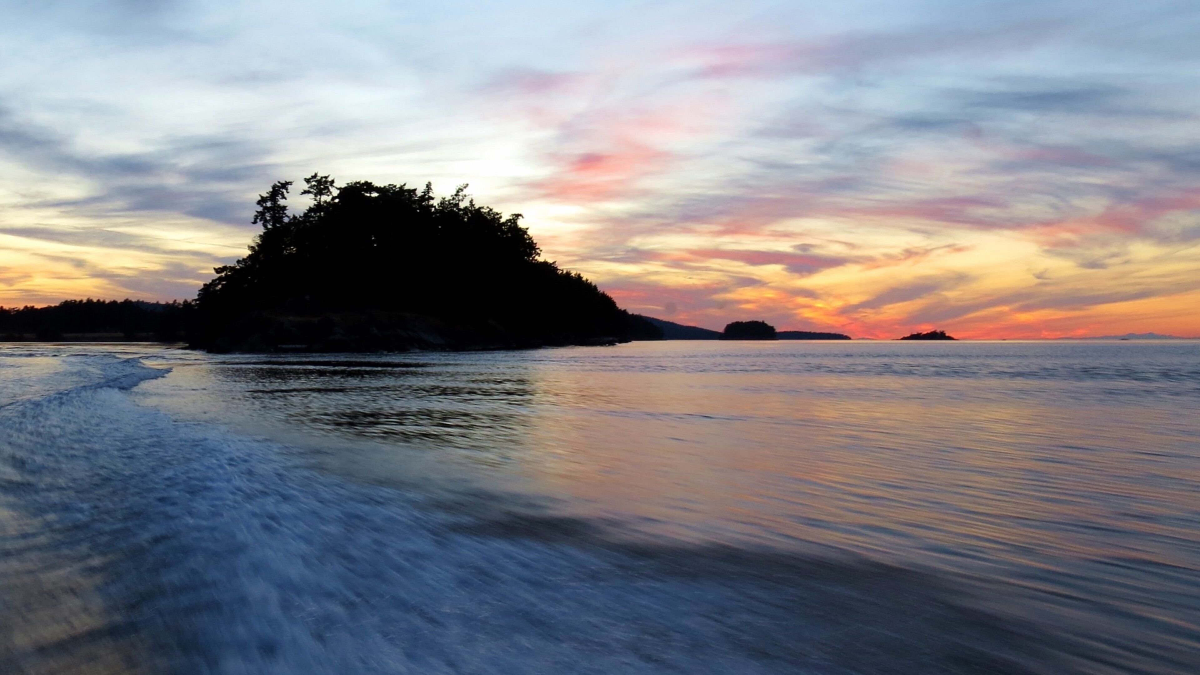 A calm sea with a small island covered in trees during sunset.