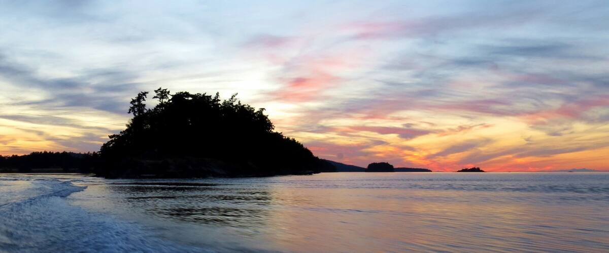 A calm sea with a small island covered in trees during sunset.
