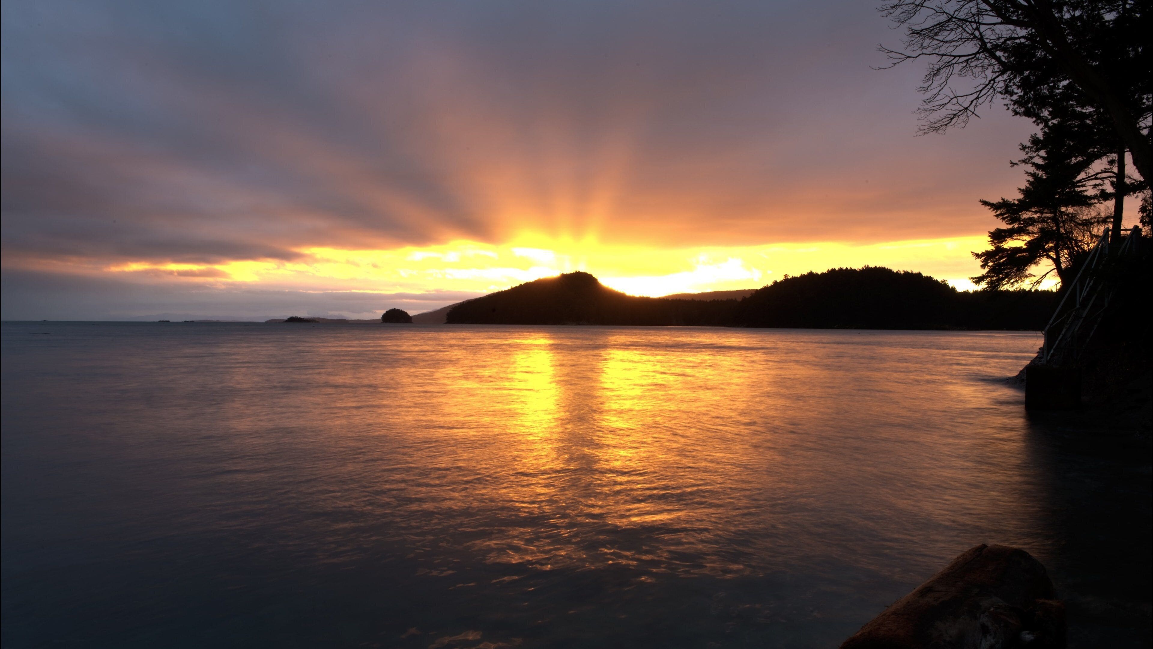 A sunset over a calm sea with a silhouette of a mountain and trees on the right.