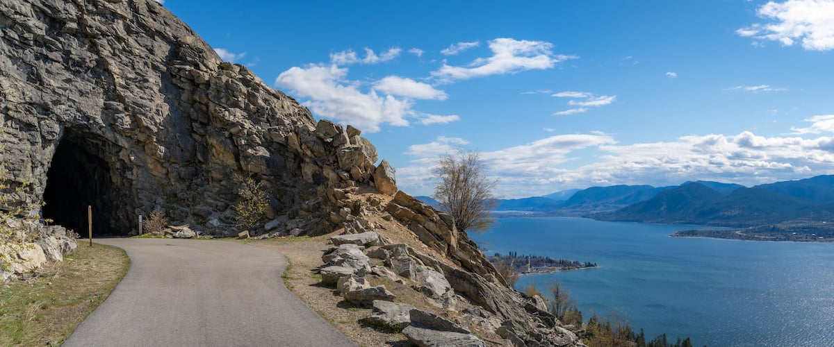 A mountain road leading to a rocky tunnel with a lake and mountains in the background.