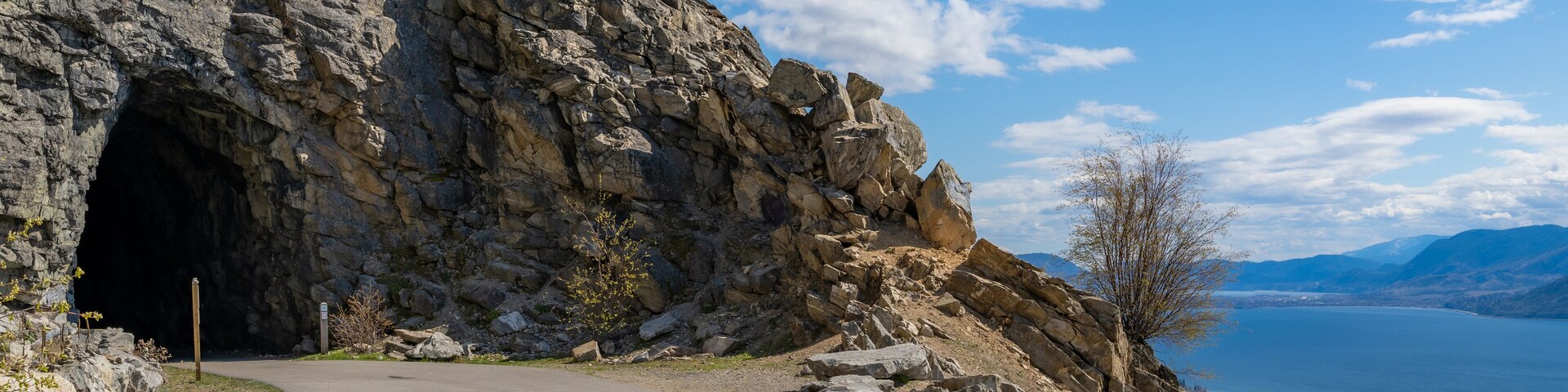 Un sendero pedregoso que lleva a un túnel, con un lago y montañas al fondo.