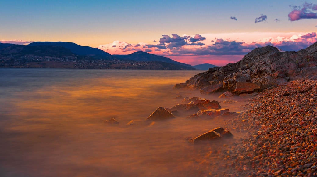 A rocky shoreline with a calm lake and distant mountains during sunset.