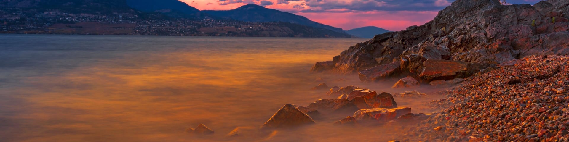 A rocky shoreline with a calm lake and distant mountains during sunset.