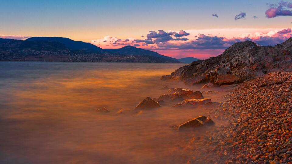 A rocky shoreline with a calm lake and distant mountains during sunset.