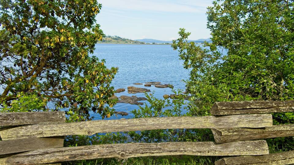 A wooden fence overlooking a calm lake with rocks in the water and trees in the foreground.