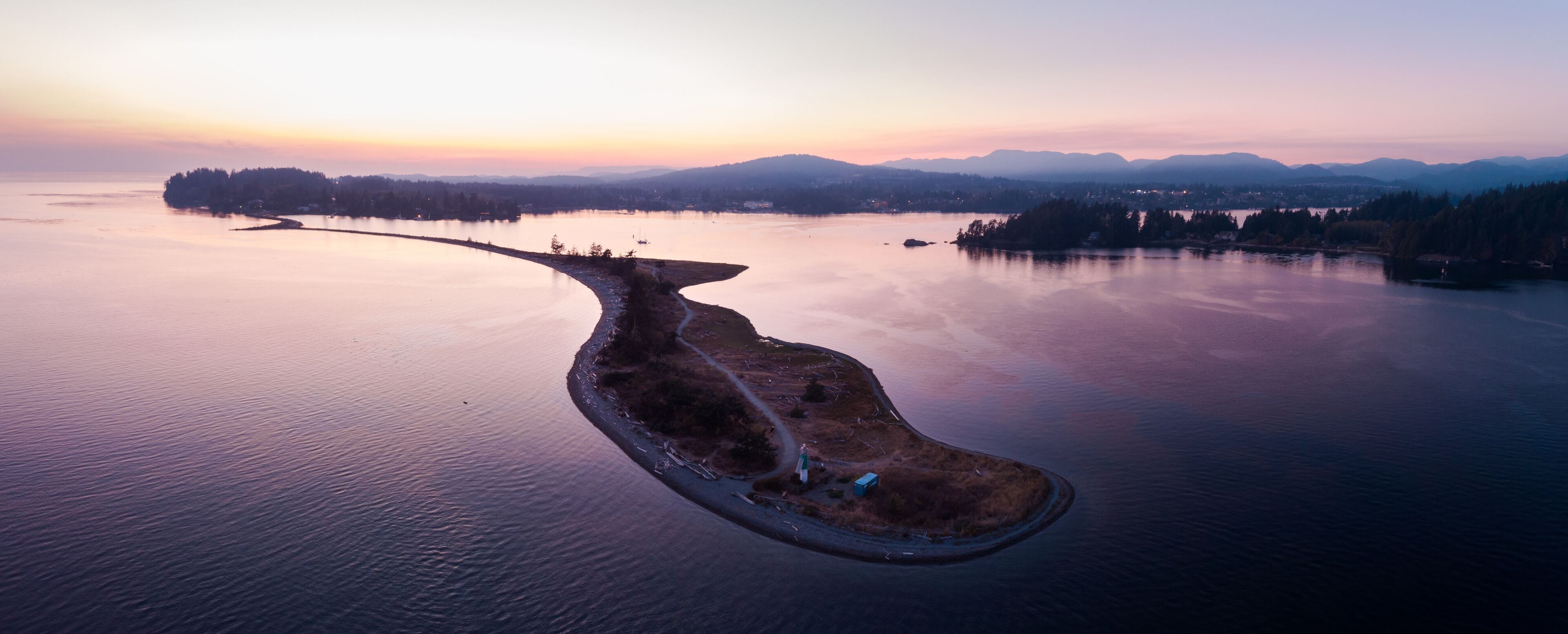 An aerial view of a peninsula with a road leading to a small island, surrounded by calm water and distant mountains.