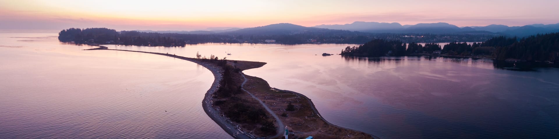 An aerial view of a peninsula with a road leading to a small island, surrounded by calm water and distant mountains.