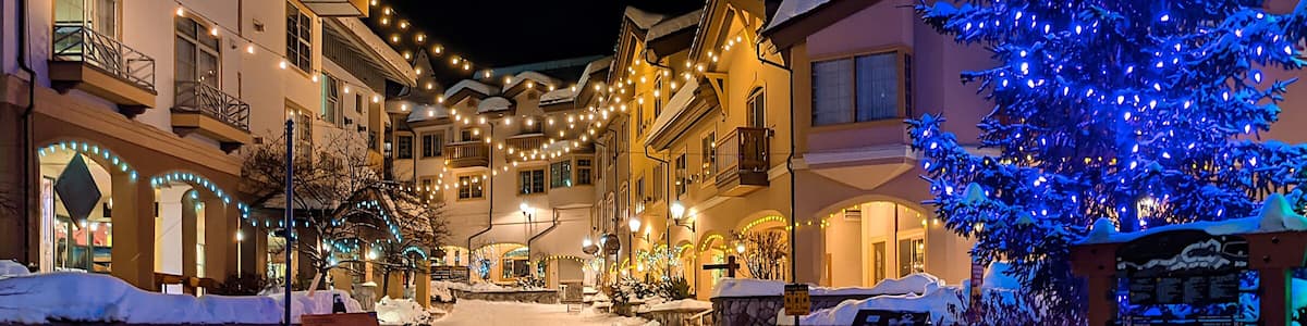 A snow-covered street lined with buildings adorned with string lights and a large Christmas tree with blue lights.