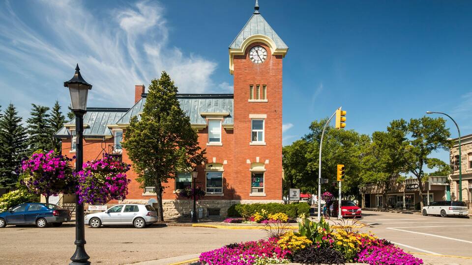 A historic brick building with a clock tower, surrounded by vibrant flower beds and a clear blue sky.