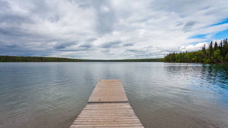 A wooden dock extending into a calm lake surrounded by dense forest under a cloudy sky.