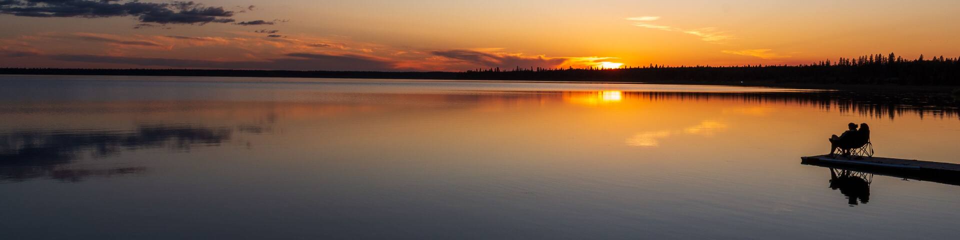 A calm lake at sunset with a clear sky, a silhouette of a dock with two people, and a forest in the background.