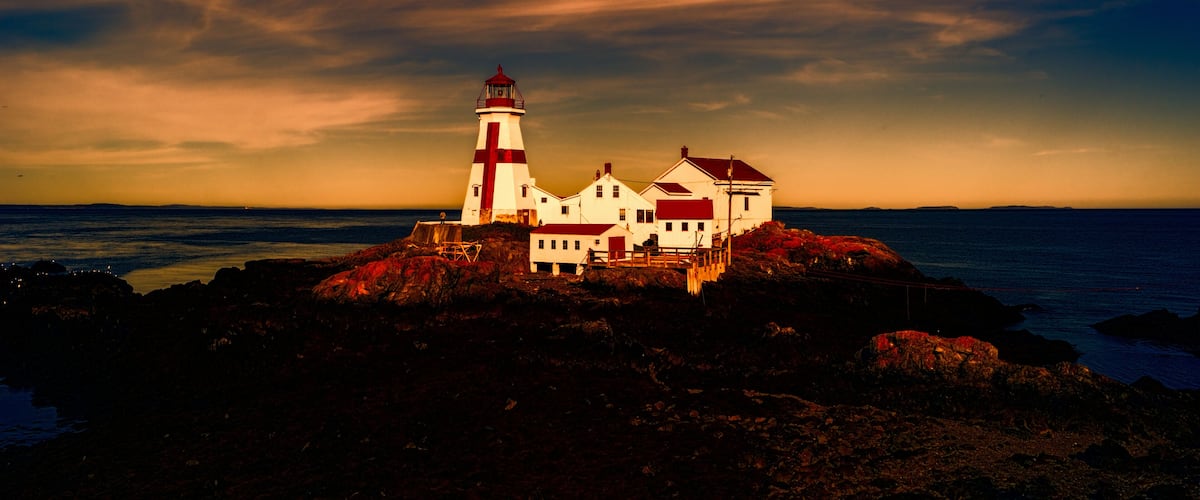 A lighthouse and white buildings on a rocky shore with the ocean in the background.