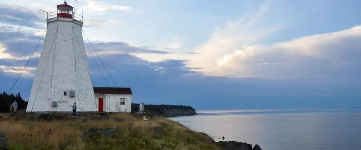 A white lighthouse with a red door on a grassy cliff overlooking calm waters.